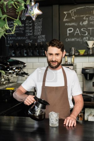 barista sonriente en delantal mirando a la cámara y sosteniendo la cafetera cerca del vidrio con cubitos de hielo en la cafetería