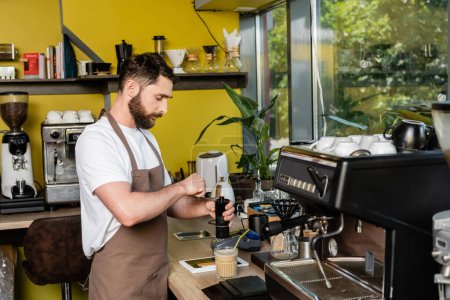 barista en delantal moliendo café cerca de la tableta digital y la bebida de café frío en la cafetería