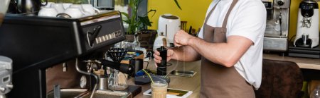 vista recortada de barista en delantal moliendo café cerca de tableta digital y café frío en la cafetería, pancarta