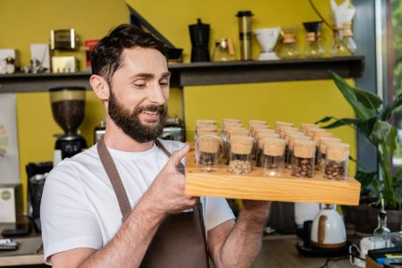 barista barbudo sonriente en delantal sosteniendo granos de café en frascos mientras trabajaba en la cafetería