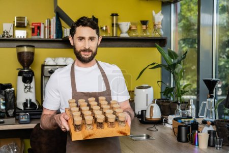 Photo for Smiling barista in apron holding jars with coffee beans and looking at camera in coffee shop - Royalty Free Image
