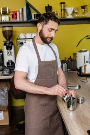 bearded barista in apron pouring coffee in metal pitcher while working in coffee shop