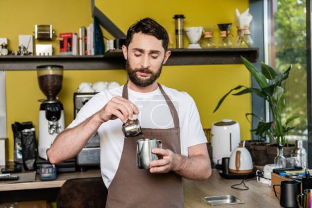 bearded barista in apron pouring coffee in metal pitcher while working in cafe on background