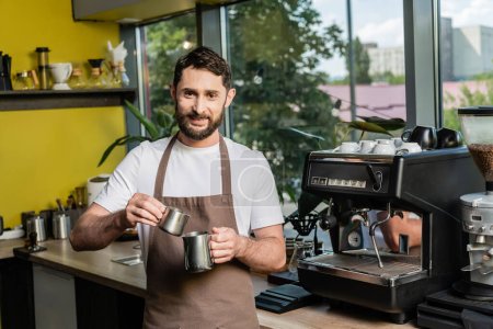 Foto de Alegre barbudo barista en delantal sosteniendo jarras cerca de la máquina de café en la cafetería - Imagen libre de derechos