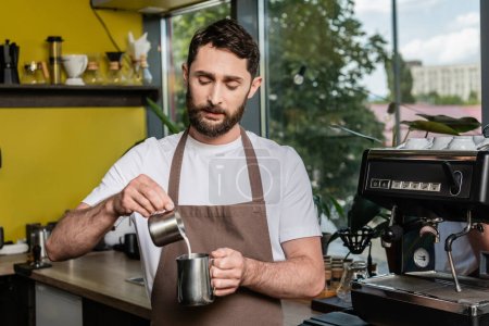 Foto de Barista barbudo enfocado en delantal sosteniendo jarras cerca de la máquina de café en la cafetería - Imagen libre de derechos