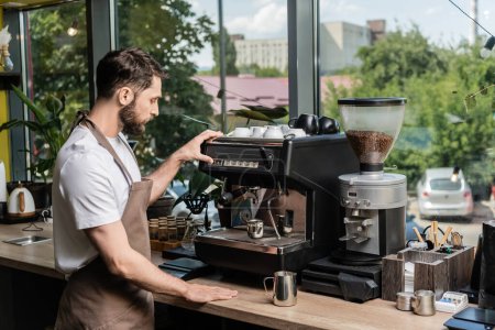 Foto de Vista lateral del barista barbudo en delantal haciendo café en la máquina de café cerca de jarras en la cafetería - Imagen libre de derechos