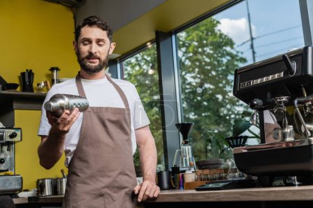 barbudo barista en delantal usando agitador mientras trabaja cerca de la máquina de café en la cafetería