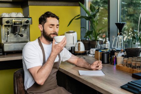 Foto de Barbudo barista en delantal oliendo café en la taza cerca de portátil en el taller en la cafetería - Imagen libre de derechos