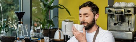 bearded barista in apron smelling coffee in cup while working in coffee shop, banner