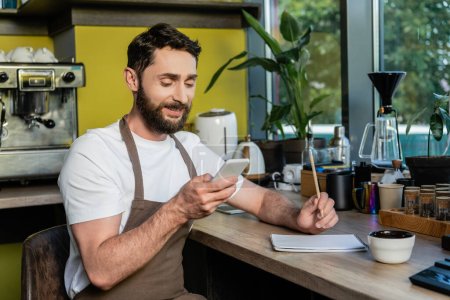 positive barista in apron using smartphone near notebook on worktop in coffee shop