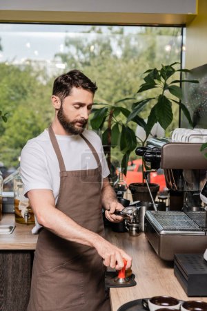 bearded barista in apron holding press and holder near coffee machine in coffee shop