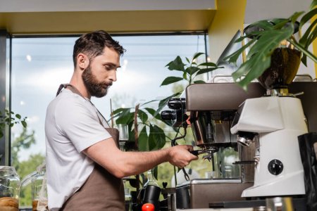 bearded barista in apron holding holder near coffee machine while working in coffee shop