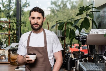 bearded barista in apron holding cup and looking at camera near coffee machine in cafe