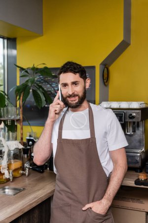 Lächelnder bärtiger Barista in Schürze, der auf dem Smartphone spricht, während er im Café im Hintergrund arbeitet