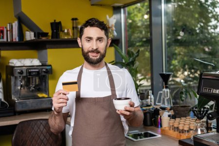 barista sonriente en delantal mirando a la cámara mientras sostiene la tarjeta de crédito y la taza en la cafetería