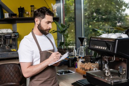 barista in apron writing on notebook while using coffee machine and working in coffee shop