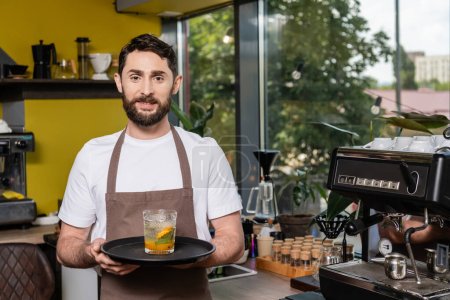 cheerful barista in apron holding cold lemonade in glass and looking at camera in coffee shop
