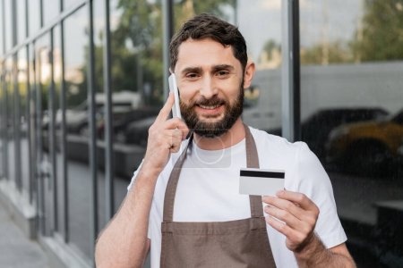 Téléchargez les photos : Gai barista tenant carte de crédit et parler sur smartphone sur la rue urbaine sur fond - en image libre de droit
