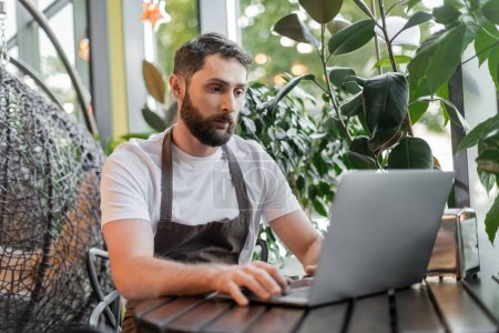 Barista in Schürze mit Laptop, während er in der Nähe von Pflanzen sitzt und im Café arbeitet