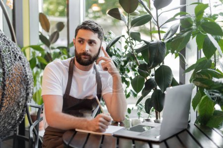 barista talking on smartphone and writing on notebook near laptop and plants in coffee shop