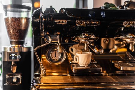Foto de Café que vierte de la máquina de café en la taza cerca de amoladora borrosa con luz solar en la cafetería - Imagen libre de derechos