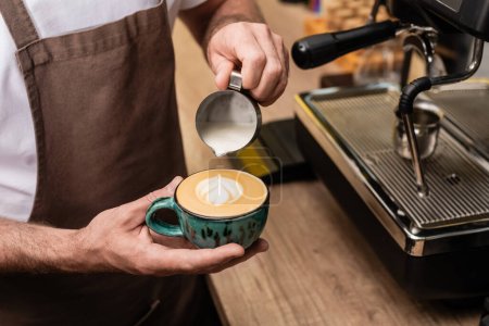 vista recortada de barista en delantal vertiendo leche y haciendo capuchino cerca de la máquina de café en la cafetería
