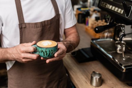 cropped view of barista in apron holding cappuccino cup while working in coffee shop
