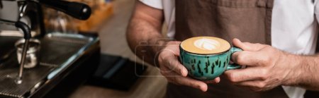 cropped view of barista in apron holding cup of cappuccino in coffee shop, banner
