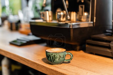 Tasse Cappuccino neben verwackelter Kaffeemaschine auf Werkstatt in Café