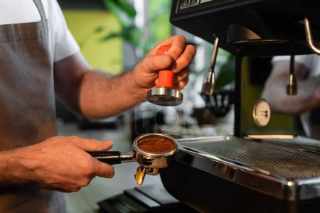 cropped view of barista in apron holding press and coffee in holder near coffee machine in cafe