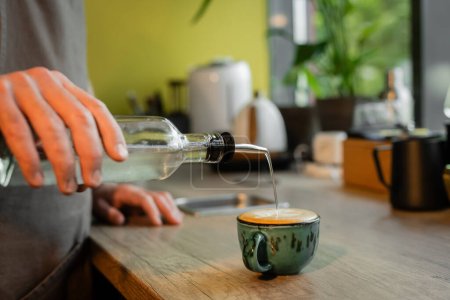 cropped view of barista pouring syrup in cup of cappuccino on worktop in coffee shop