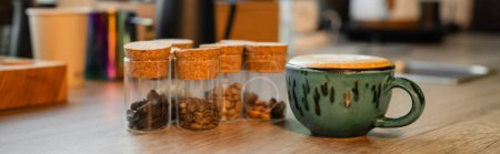 cup of cappuccino near coffee beans in jars on wooden worktop in blurred coffee shop, banner