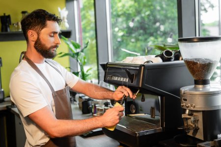 barbudo barista limpieza de la máquina de café boquilla con trapo mientras trabaja en la cafetería