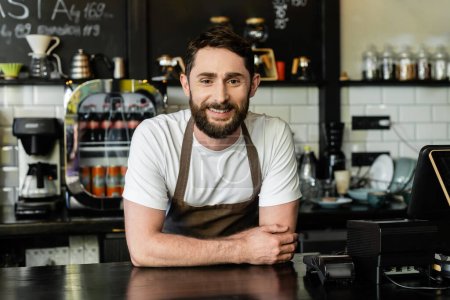 Photo for Cheerful bearded barista looking at camera while standing near bar in coffee shop - Royalty Free Image
