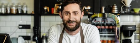 portrait of positive and bearded barista in apron looking at camera in coffee shop, banner