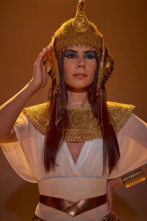 Porträt einer brünetten Frau im ägyptischen Outfit, die die Hand auf der Hüfte hält, auf braunem Hintergrund mit Schatten