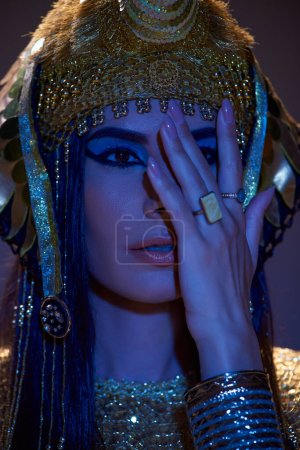 Portrait of woman in Egyptian headdress covering face and posing on brown background with blue light