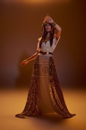 Longitud completa de la mujer con estilo en aspecto egipcio y tocado posando en luz sobre fondo marrón
