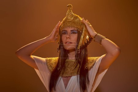 Beautiful brunette woman in Egyptian outfit touching headdress in light on brown background