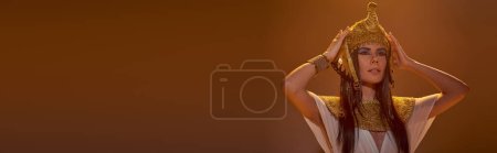Fashionable woman in Egyptian attire touching headdress in lighting on brown background, banner