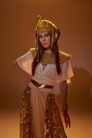 Modelo morena con estilo en aspecto egipcio y maquillaje audaz posando y de pie sobre fondo marrón