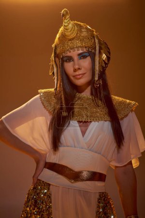 Brunette woman in ancient Egyptian outfit and headdress looking at camera isolated on brown