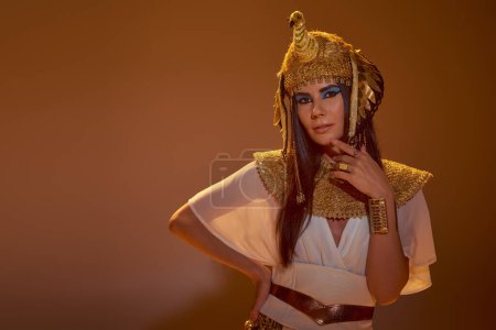 Stilvolle Frau in ägyptischer Kleidung und Kopfbedeckung berührt Kinn und posiert auf braunem Hintergrund