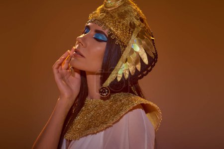 Porträt einer Frau mit kühnem Make-up und ägyptischer Kopfbedeckung, die die Wange berührt, isoliert auf braun
