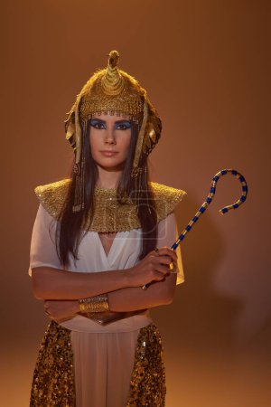 Brunette woman in egyptian attire and headdress holding crook and standing on brown background