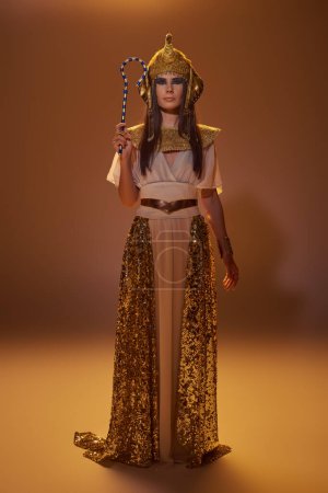 Full length of stylish woman in egyptian attire holding crook and standing on brown background