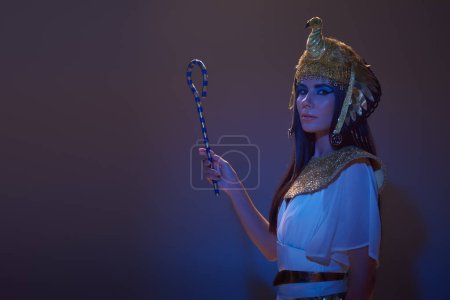 Brünette Frau in ägyptischer Kleidung und Kopfbedeckung mit Gauner auf braunem Hintergrund mit blauem Licht
