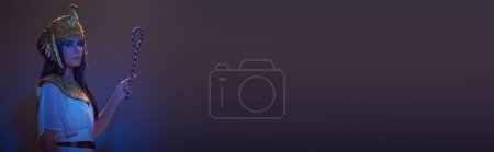 Téléchargez les photos : Femme dans le regard égyptien tenant escroc et regardant la caméra sur brun avec éclairage bleu, bannière - en image libre de droit