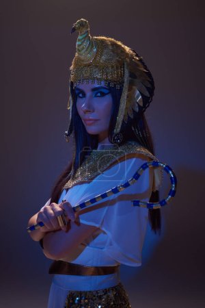 Portrait of woman in egyptian look holding crook and posing on brown background with blue light