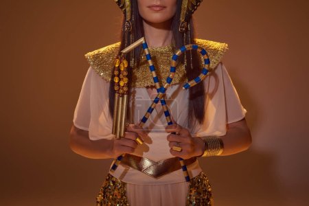 Foto de Vista recortada de la mujer en mirada egipcia sosteniendo ladrón y flail sobre fondo marrón - Imagen libre de derechos